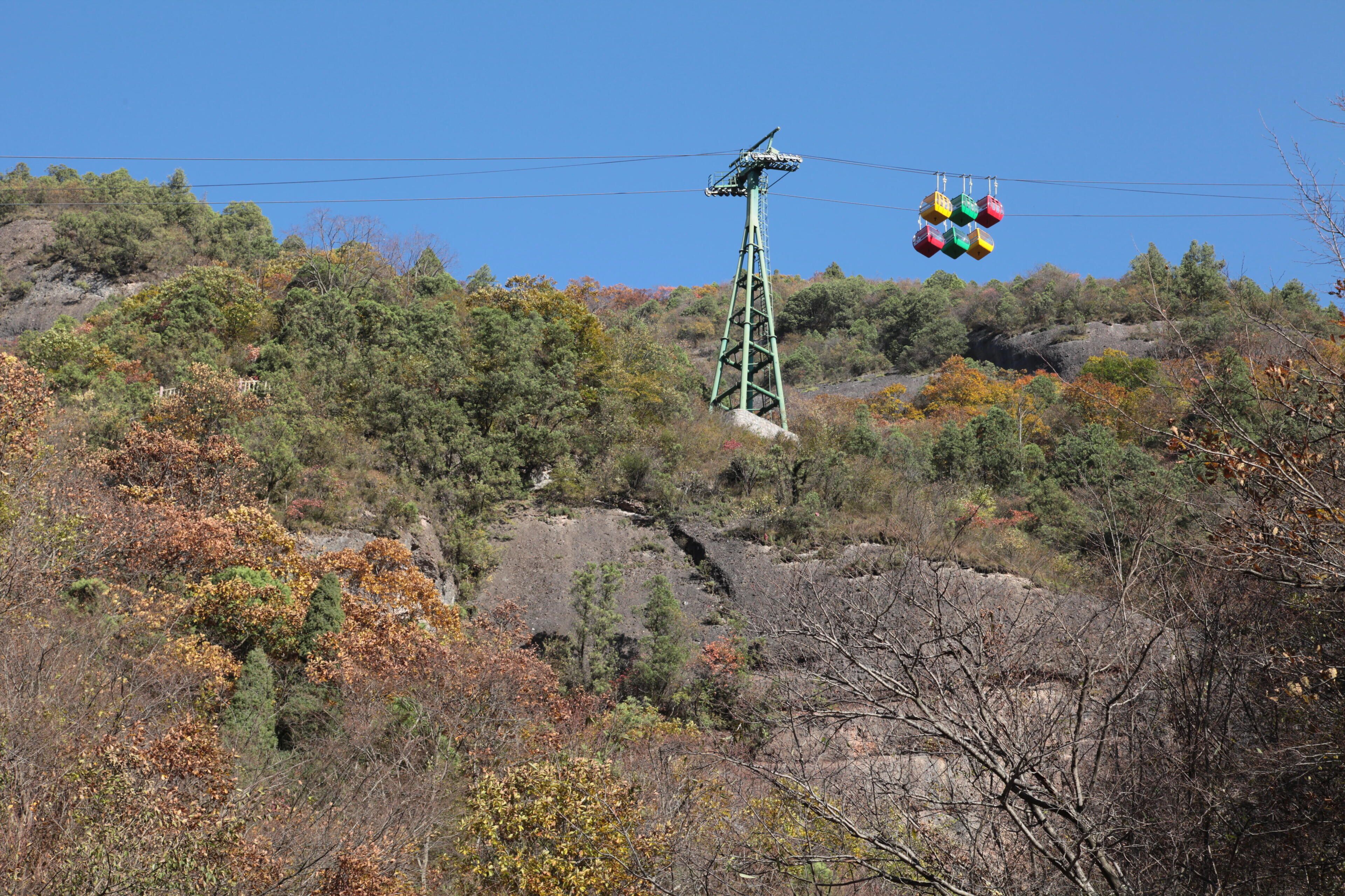 A cable car system with colorful gondolas against a backdrop of autumn-colored trees and rocky terrain.
