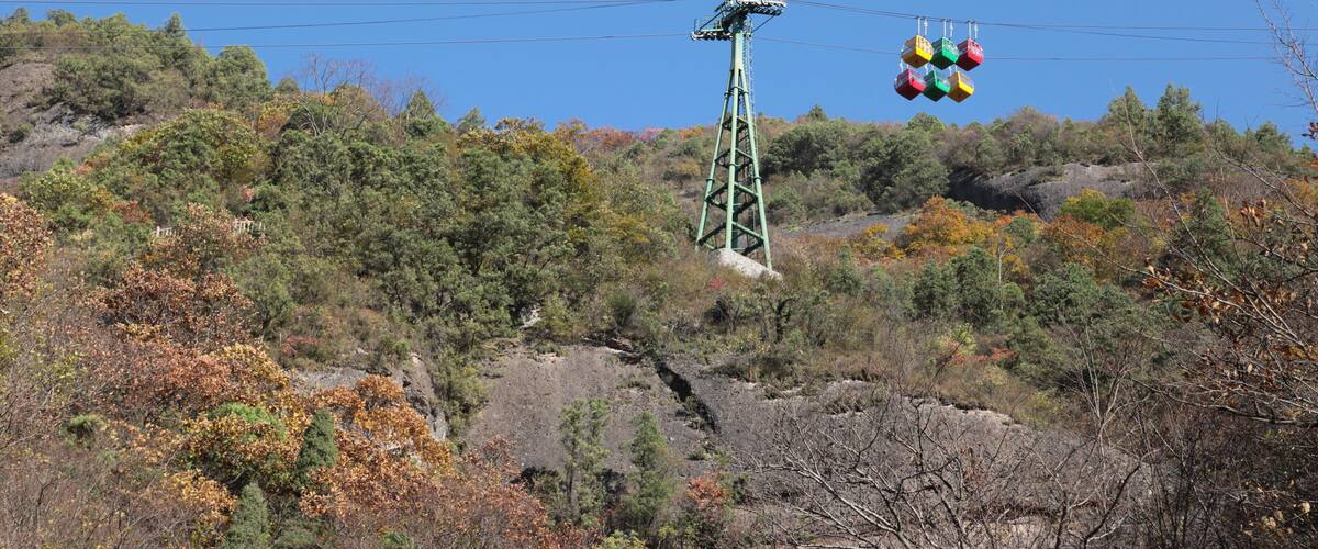 A cable car system with colorful gondolas against a backdrop of autumn-colored trees and rocky terrain.
