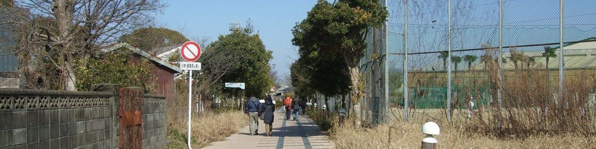 Un sendero pavimentado con un cartel de 'prohibido el paso', una valla y personas caminando.
