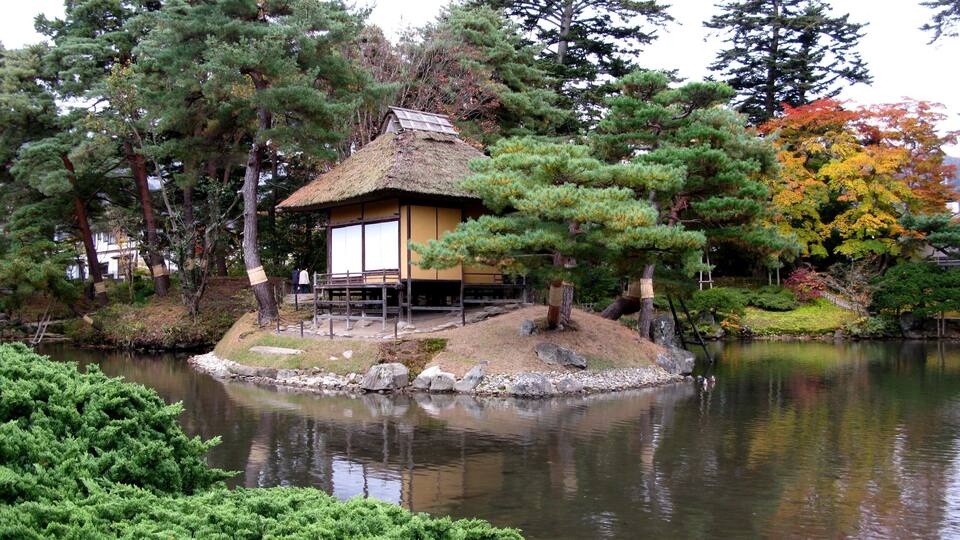 A traditional Japanese-style house on an island surrounded by a pond, with trees and autumn foliage in the background.