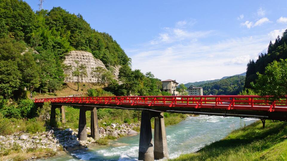 A red bridge over a river with a rocky riverbed, surrounded by lush greenery and hills.