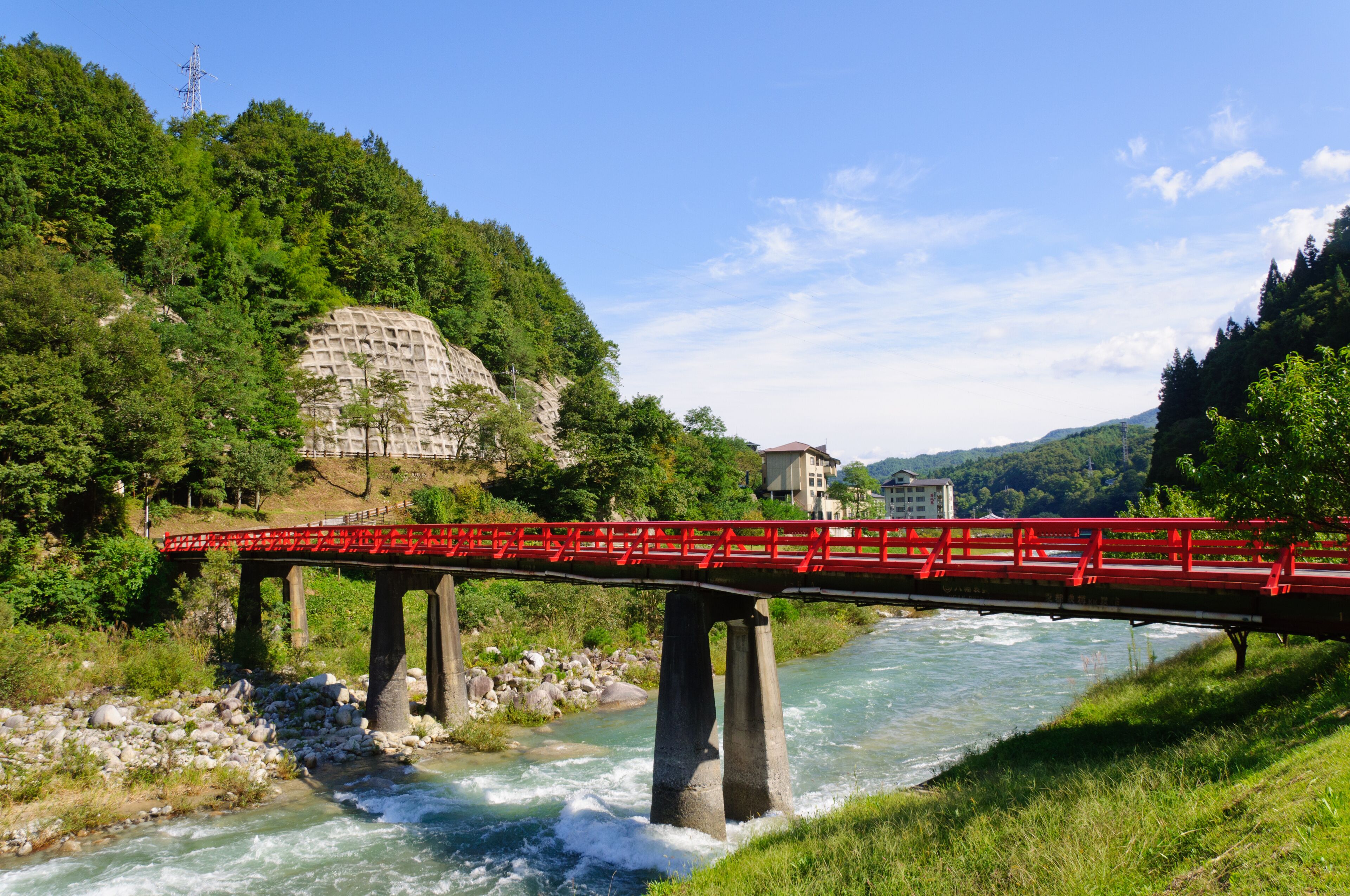 A red bridge over a river with a rocky riverbed, surrounded by lush greenery and hills.