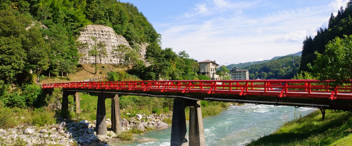 A red bridge over a river with a rocky riverbed, surrounded by lush greenery and hills.