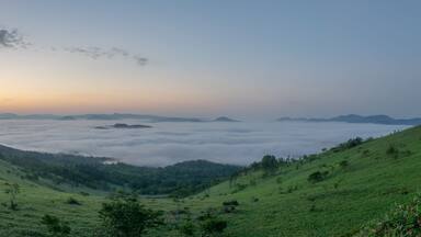 A valley with a road curving through rolling hills, a dense fog layer, and distant mountains under a clear sky.