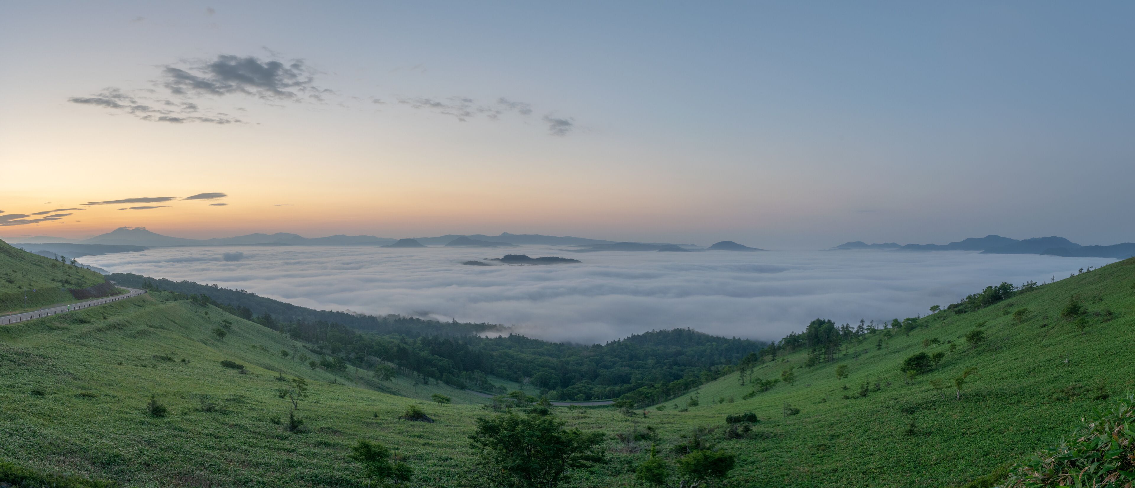 A valley with a road curving through rolling hills, a dense fog layer, and distant mountains under a clear sky.