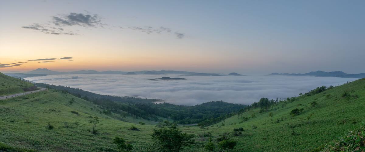 A valley with a road curving through rolling hills, a dense fog layer, and distant mountains under a clear sky.