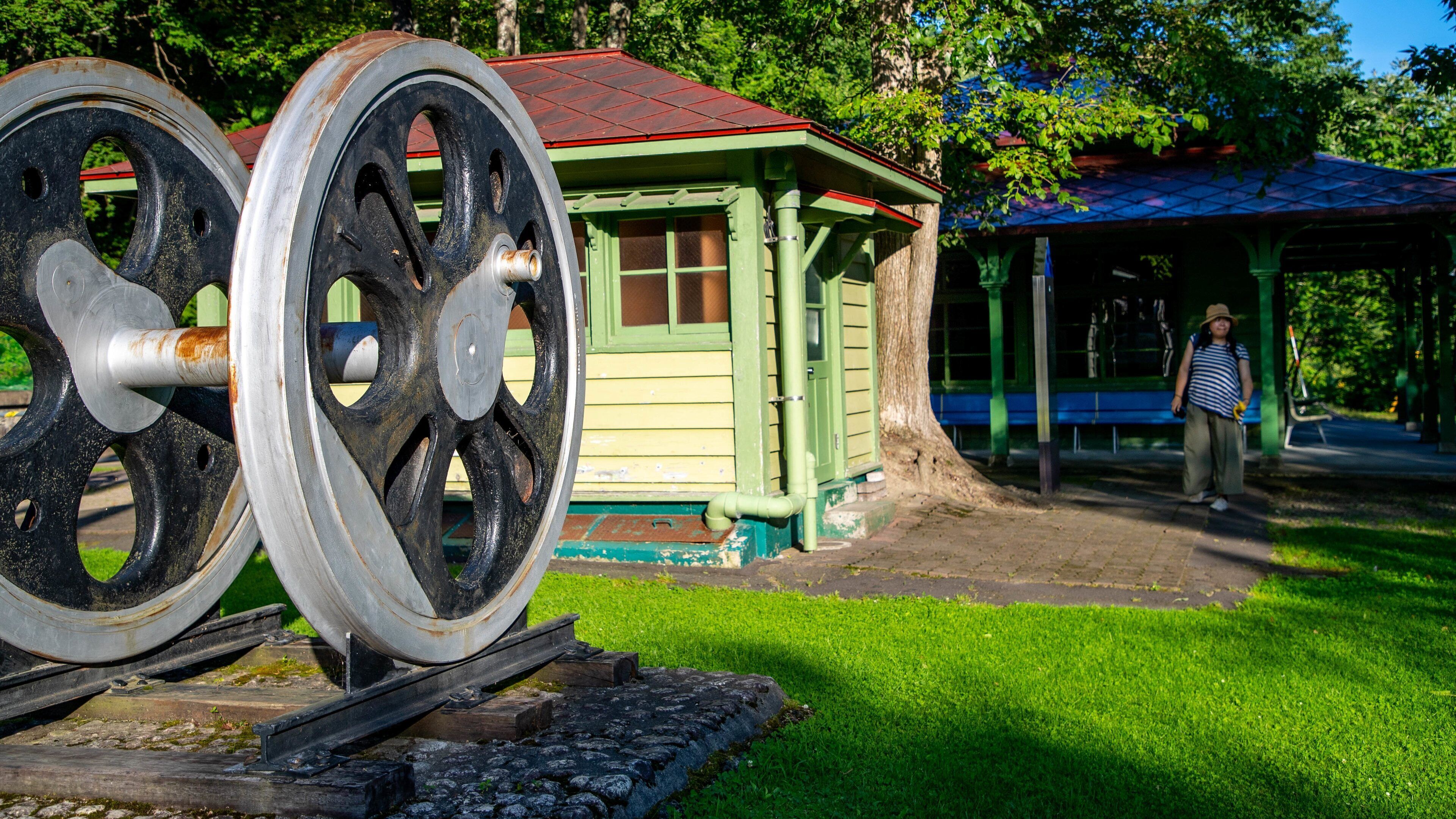 A historical train wheel, a green wooden structure, and a covered bench area.