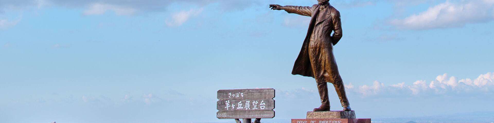A statue of a man pointing to the right, with a cityscape in the background and a clear sky.