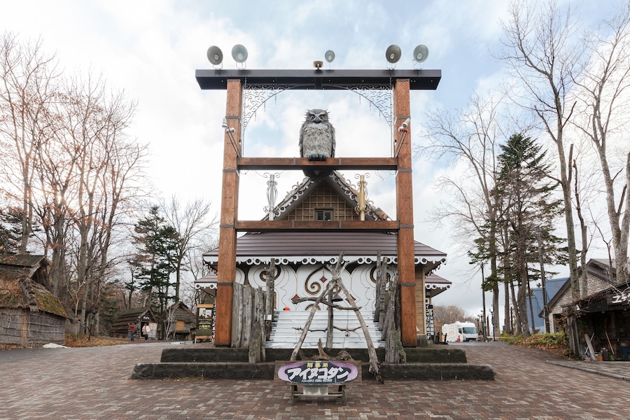 A traditional Japanese gate with an owl sculpture, surrounded by trees and a brick pathway.