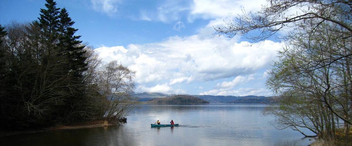 A calm lake with two people in a canoe, surrounded by trees and distant mountains.