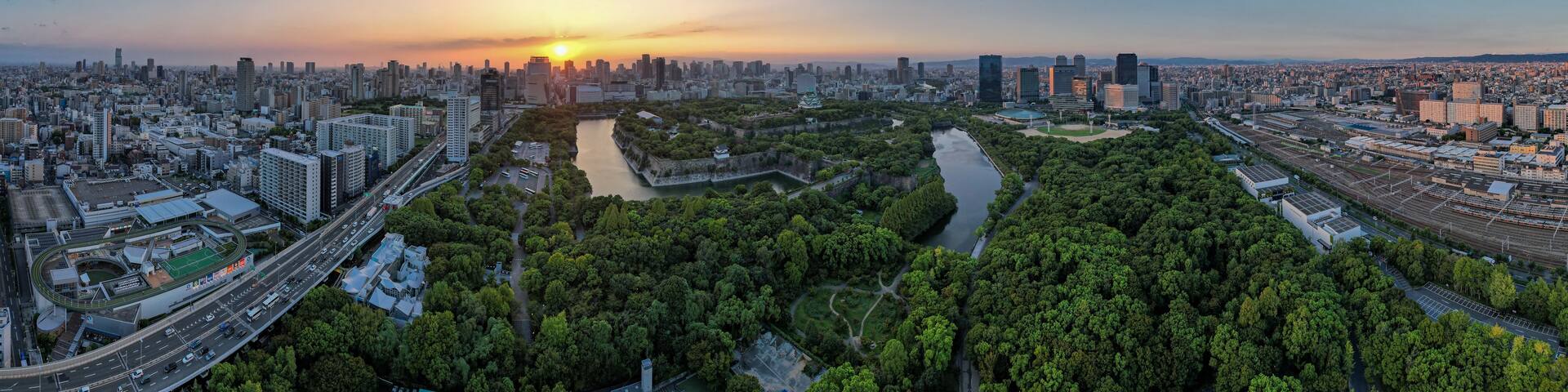 A cityscape at sunset with a river running through the center, surrounded by greenery and modern buildings.