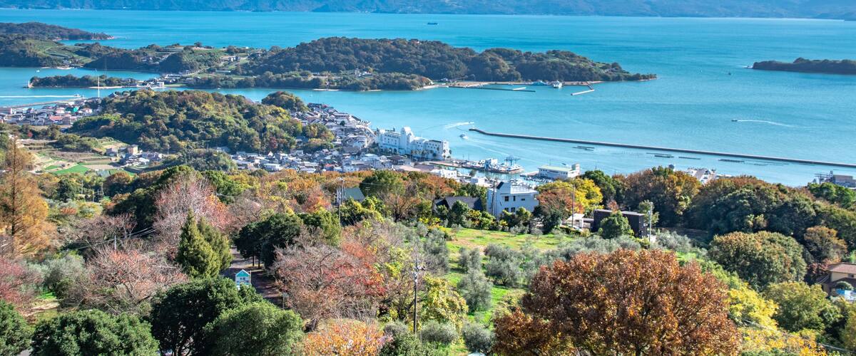 A coastal town with a harbor, mountains in the background, and autumn foliage.