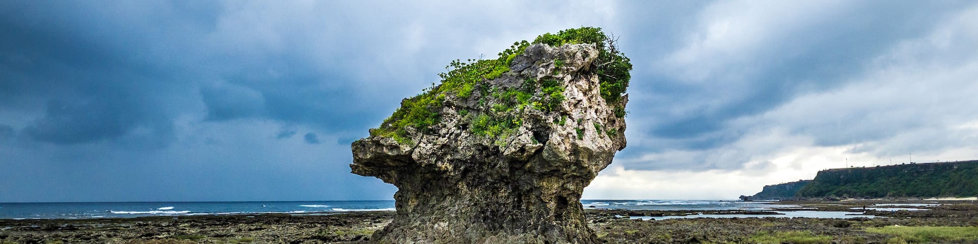 A prominent rock formation with a small island on top, surrounded by a coastal landscape with grassy areas and a body of water.