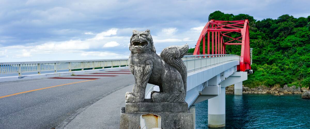 A stone lion statue on a bridge with a red truss structure and a body of water in the background.