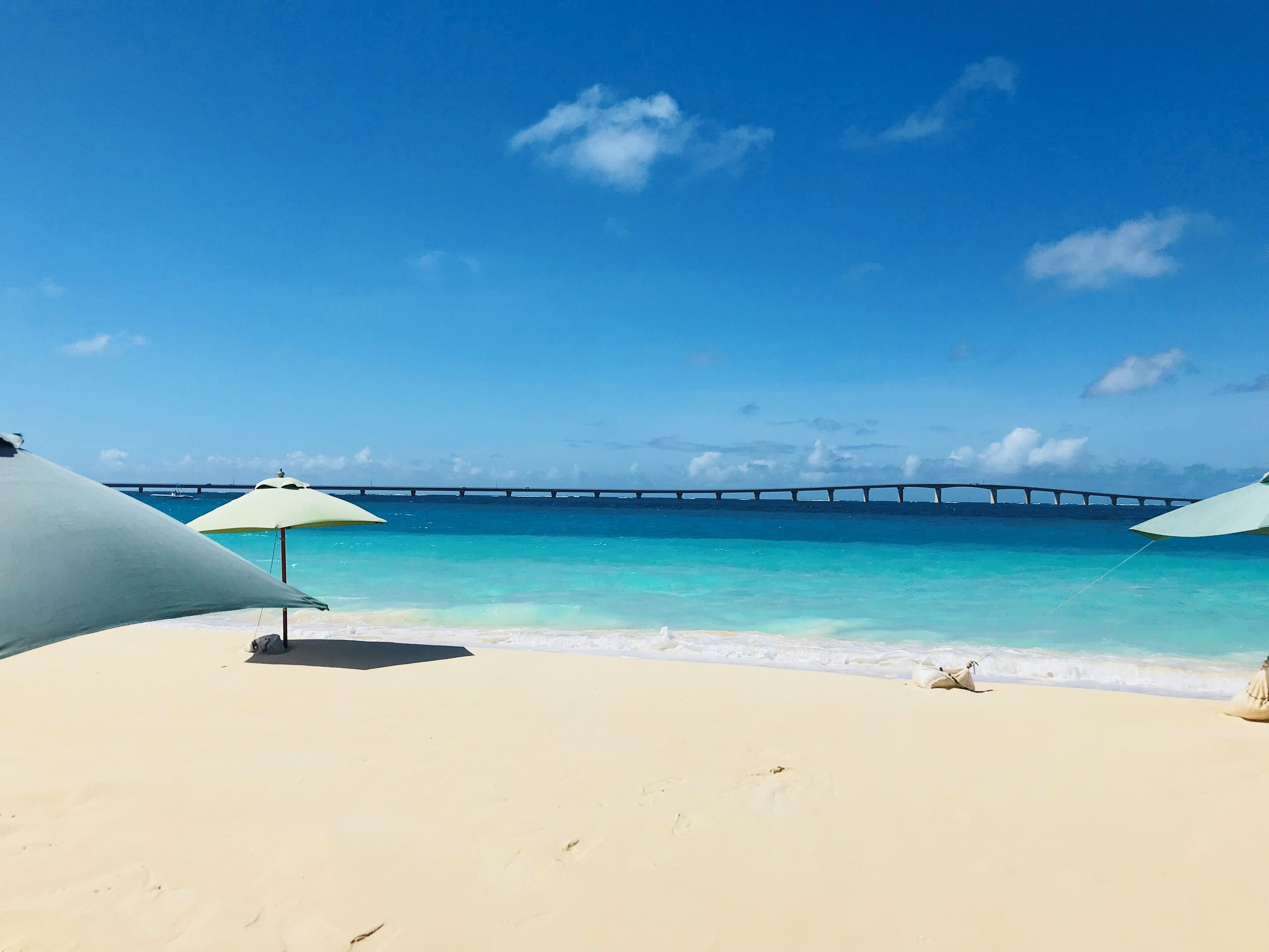 A beach with white sand and turquoise water, two beach umbrellas, and a bridge in the background.