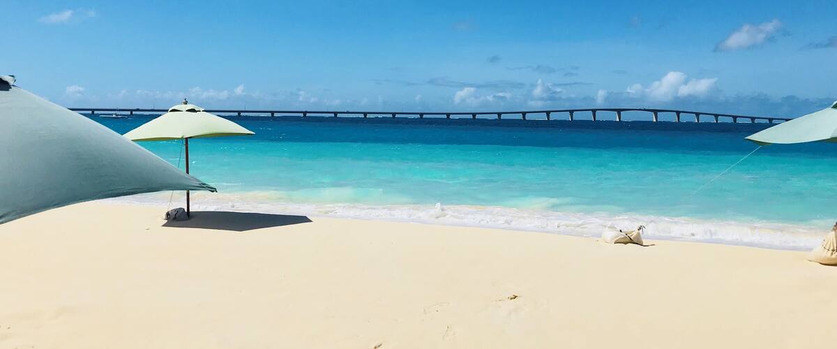 A beach with white sand and turquoise water, two beach umbrellas, and a bridge in the background.