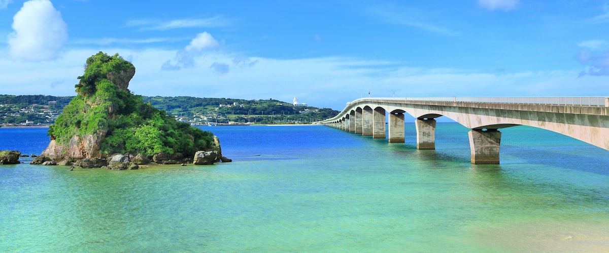 Un paisaje costero con un puente sobre aguas azules cristalinas, un promontorio rocoso y una costa distante con edificios.