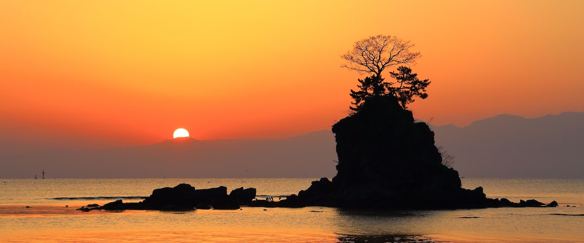 Ein Sonnenuntergang über ruhigem Meer mit einer markanten Felsformation im Vordergrund und einem Baum darauf.