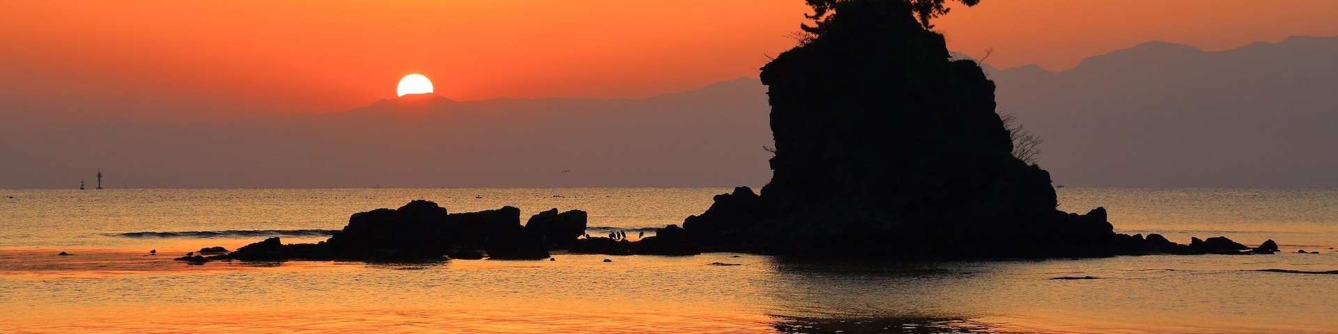 Um pôr do sol sobre um mar calmo, com uma rocha grande em primeiro plano e uma árvore no topo.