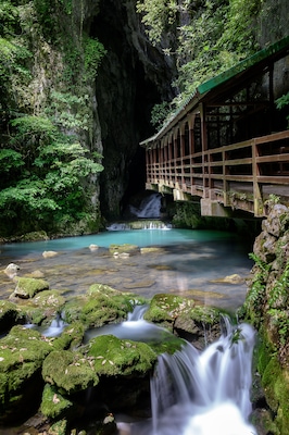 Een houten brug over een heldere beek met mosbedekte rotsen en een waterval op de achtergrond.