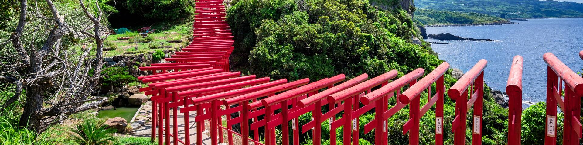 A red wooden pathway leading up a hill with lush greenery and a view of the sea.