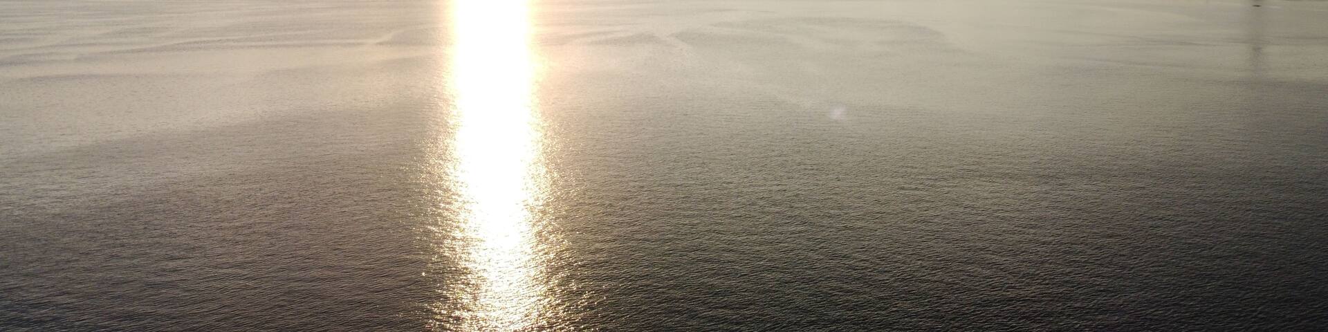 A calm body of water reflecting the sun, with mountains in the background and industrial structures on the right.