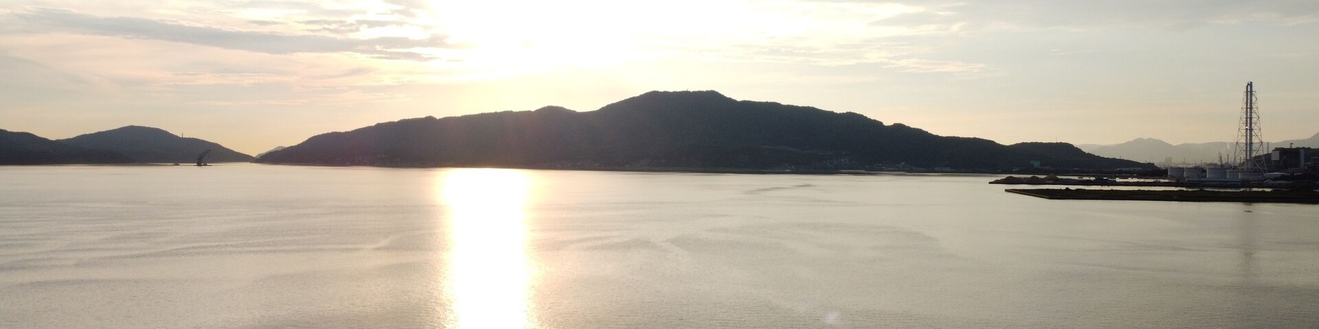 Água calma refletindo o sol, com montanhas ao fundo e estruturas industriais à direita.