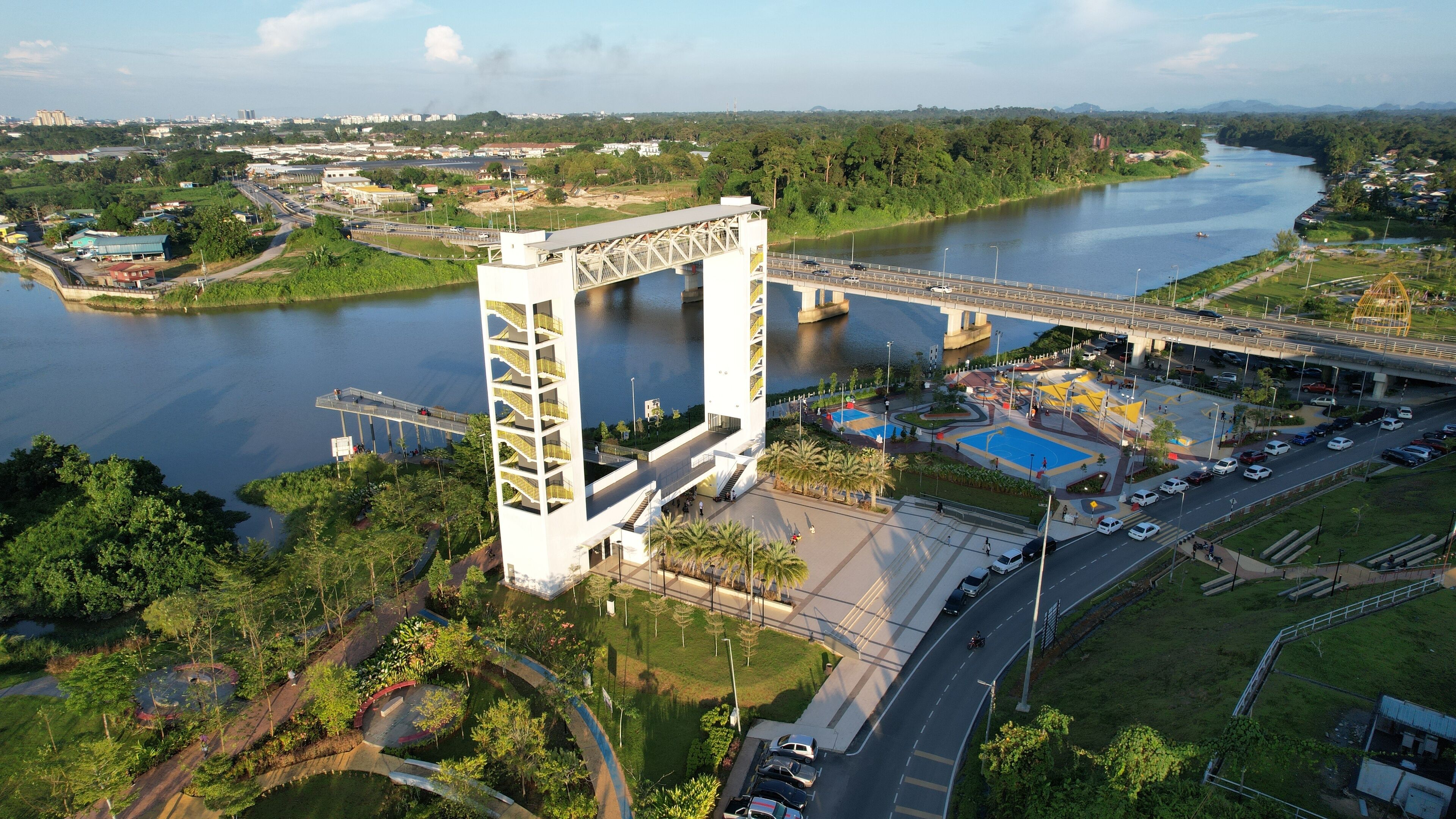 A large white bridge with a unique arch design spanning a river, surrounded by greenery and urban development.