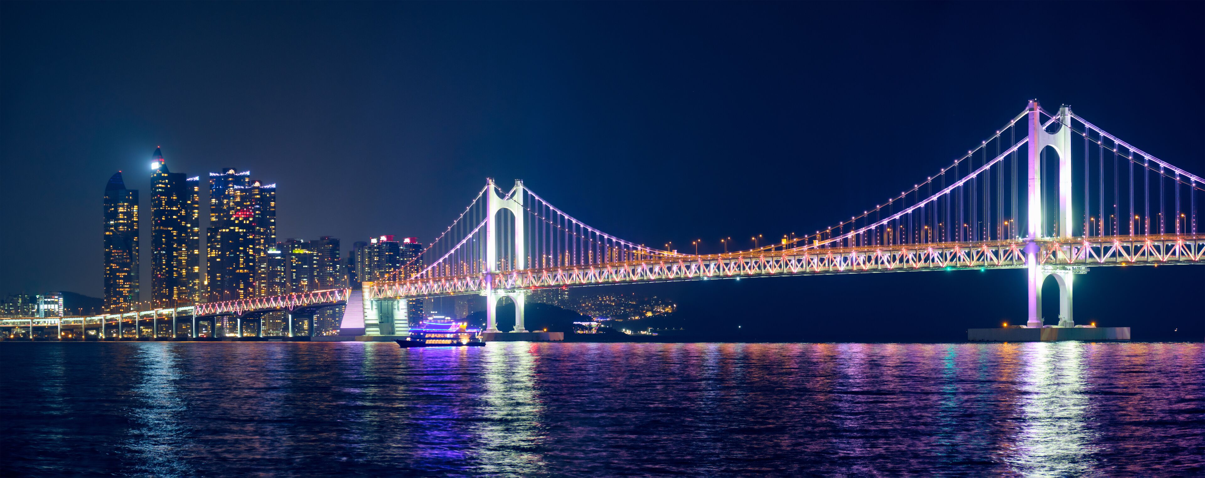 A brightly lit suspension bridge at night with city buildings in the background.