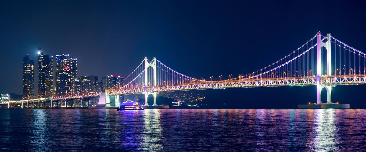 A brightly lit suspension bridge at night with city buildings in the background.