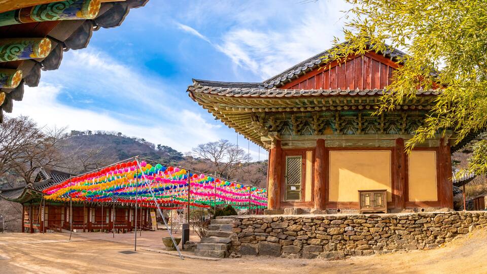 A traditional Korean pavilion with colorful lanterns, a stone wall, and a mountainous backdrop.