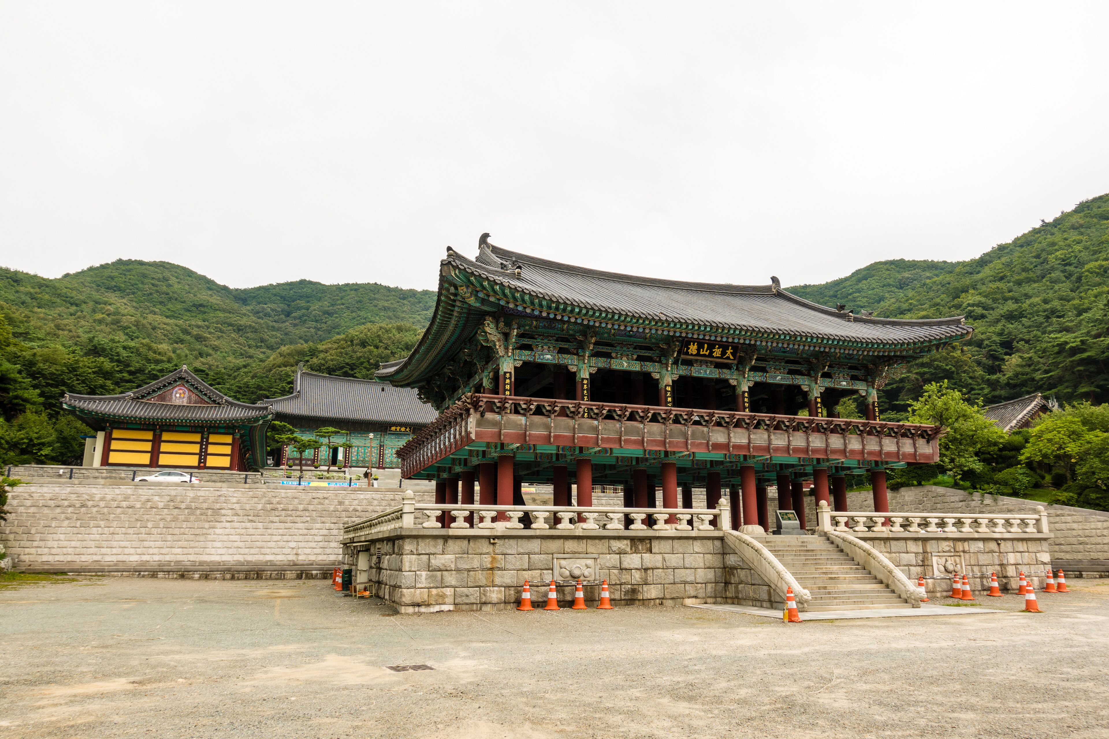 A traditional Korean building with a curved roof and red pillars, surrounded by a stone wall and set against a backdrop of green hills.