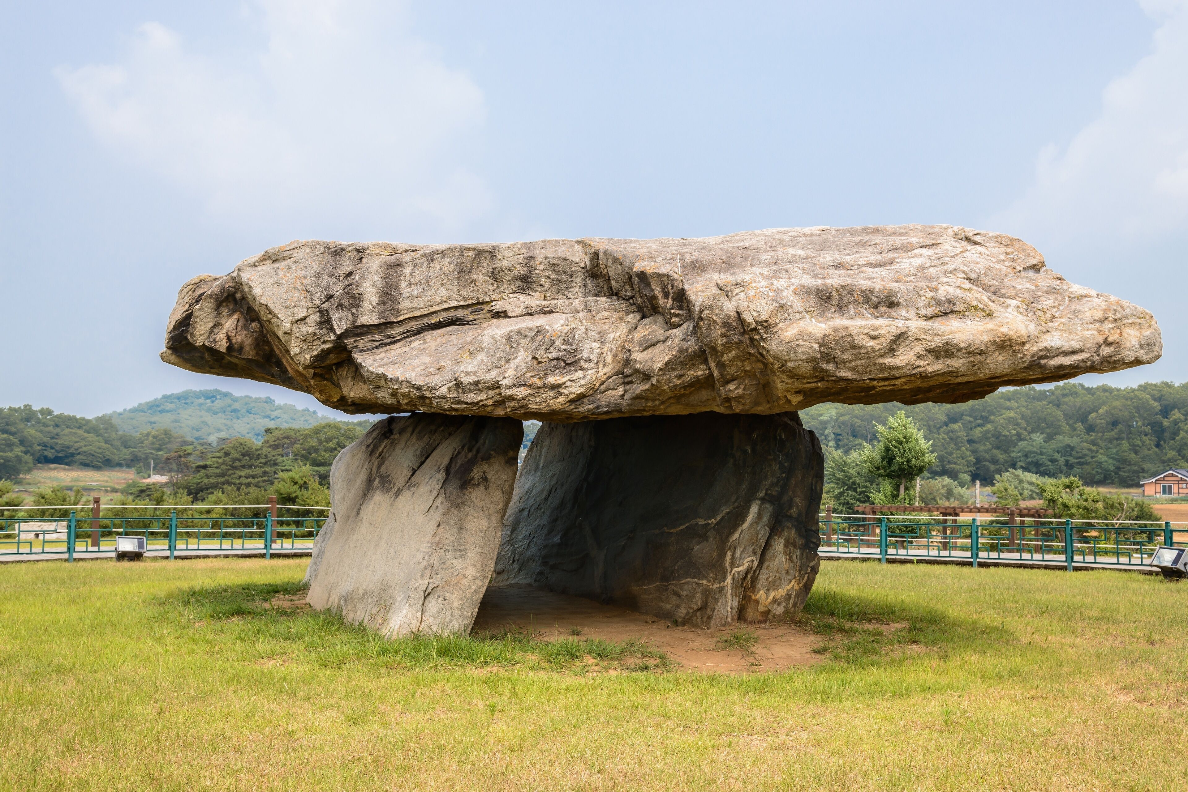 A large rock formation balanced on a smaller base, surrounded by grassy fields and a fence.