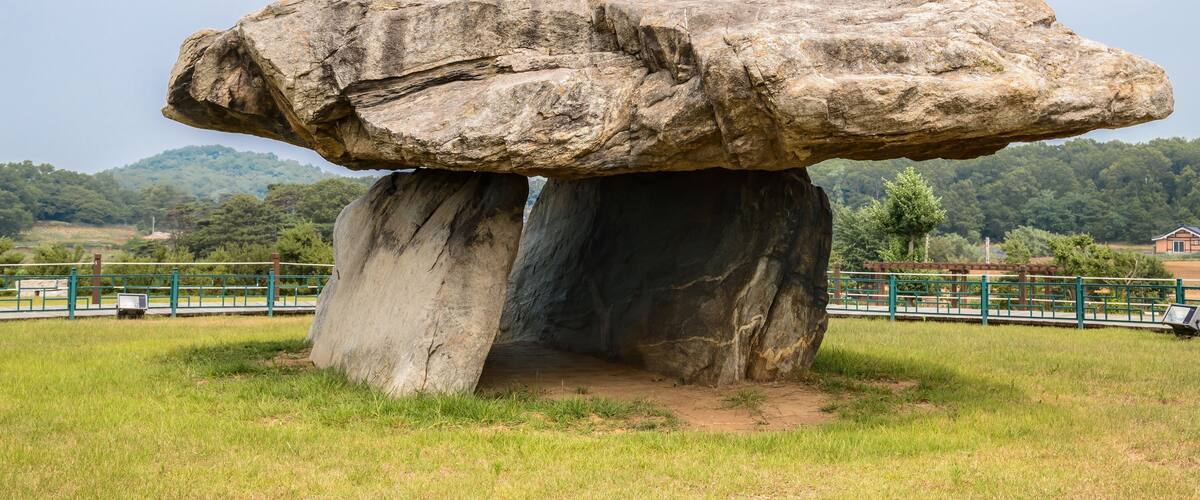A large rock formation balanced on a smaller base, surrounded by grassy fields and a fence.