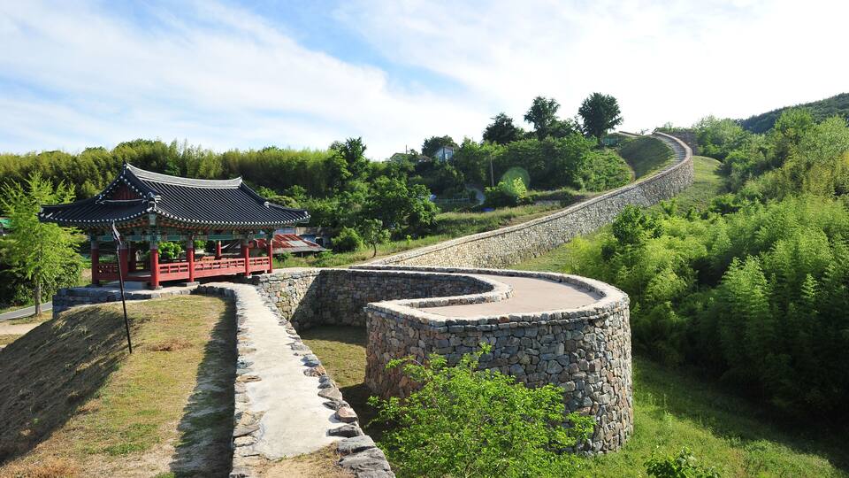 A traditional Korean pavilion, a stone wall, and a curved pathway in a lush green landscape.