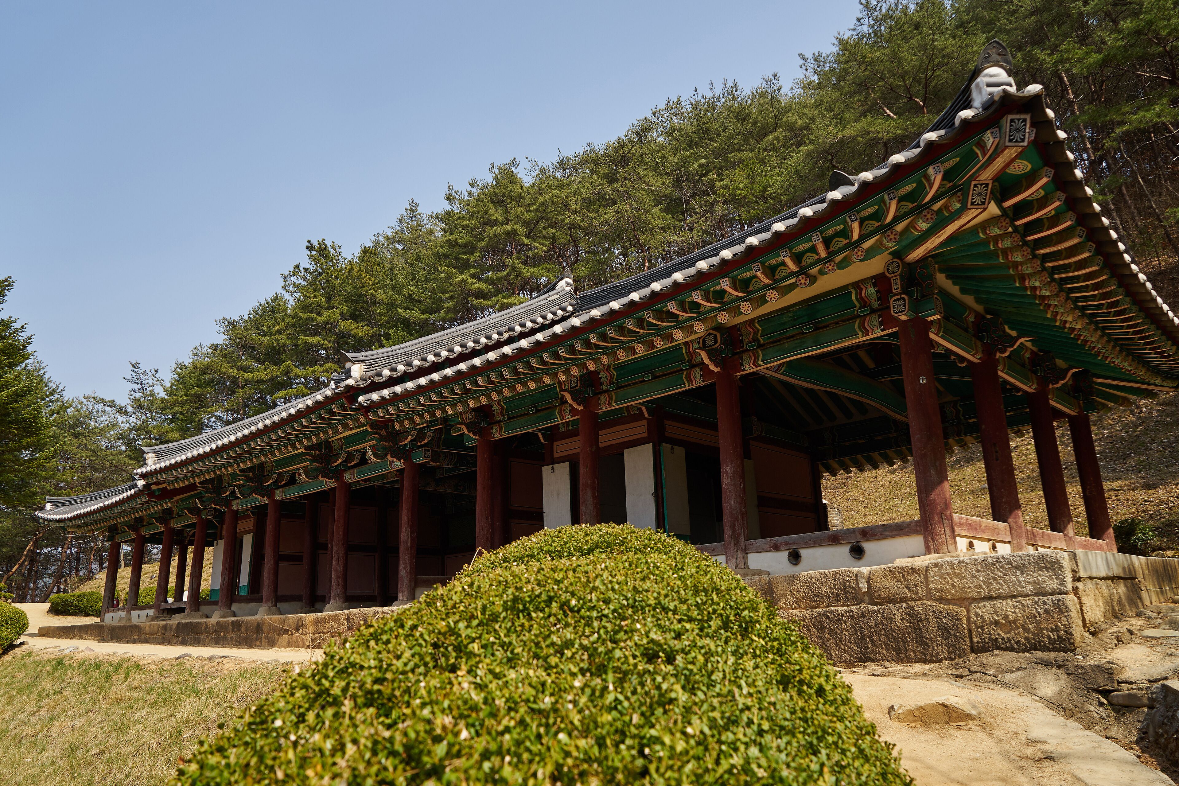 A traditional Korean pavilion with ornate roof tiles and wooden pillars, surrounded by lush greenery and trees.