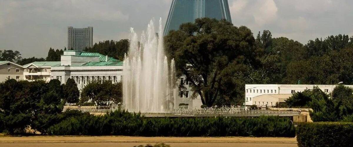 A tall, pointed building with a green roof, surrounded by trees and a fountain.