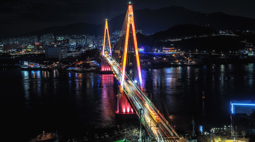 Un puente bien iluminado con luces de colores que se reflejan en el agua, un paisaje urbano al fondo y montañas a lo lejos.