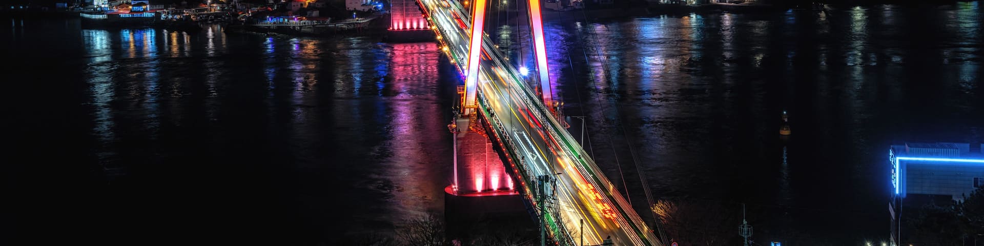 A brightly lit bridge with colorful lights reflecting on the water, a cityscape in the background, and mountains in the distance.