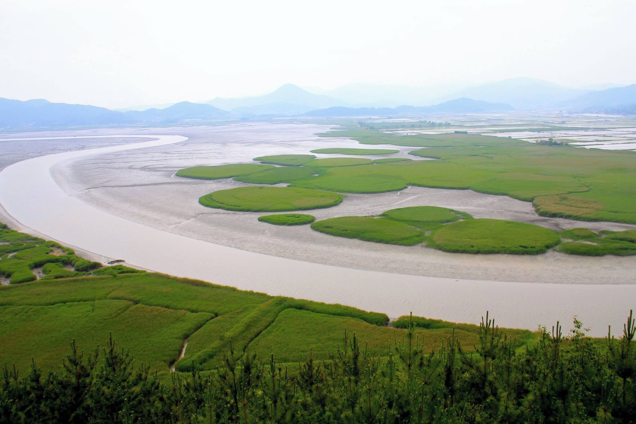 A river meandering through green fields with circular water bodies and distant mountains.