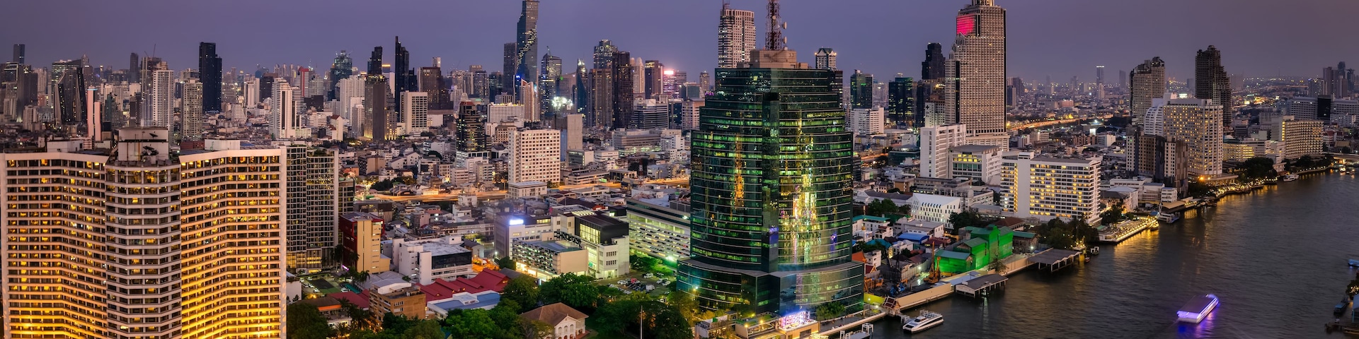 Paisagem urbana ao anoitecer, com edifícios iluminados, um rio e uma ponte.