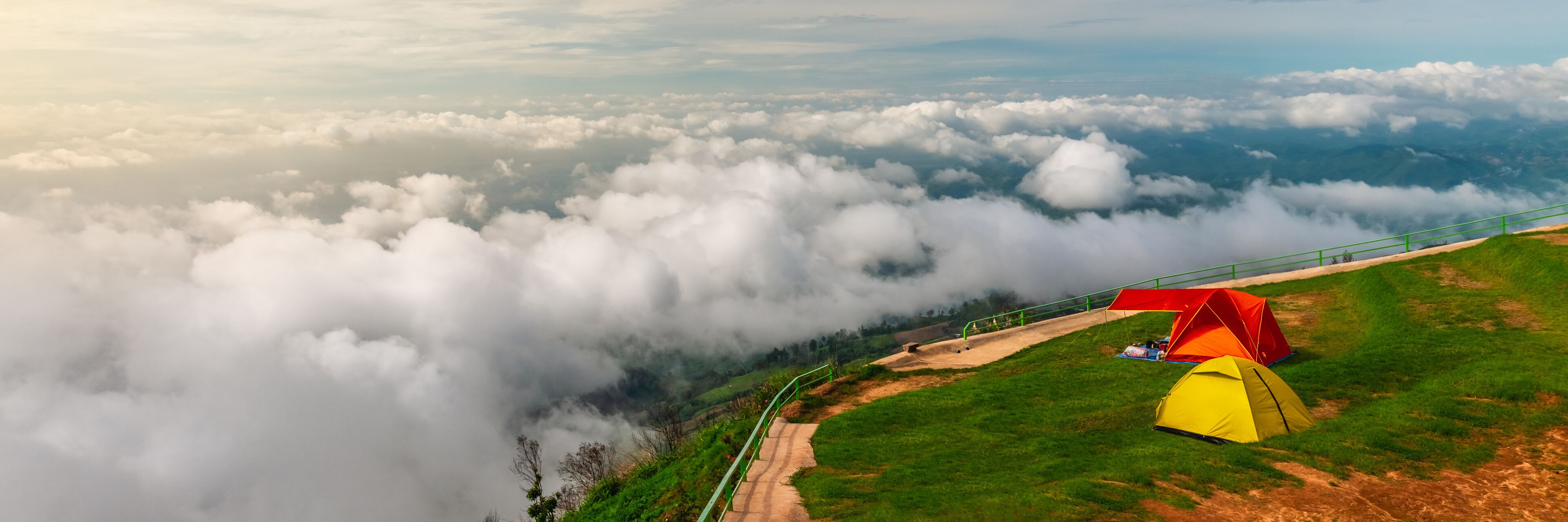 A mountainous landscape with a red and yellow tent on a grassy slope, surrounded by clouds.