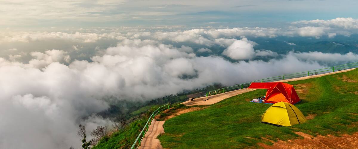A mountainous landscape with a red and yellow tent on a grassy slope, surrounded by clouds.