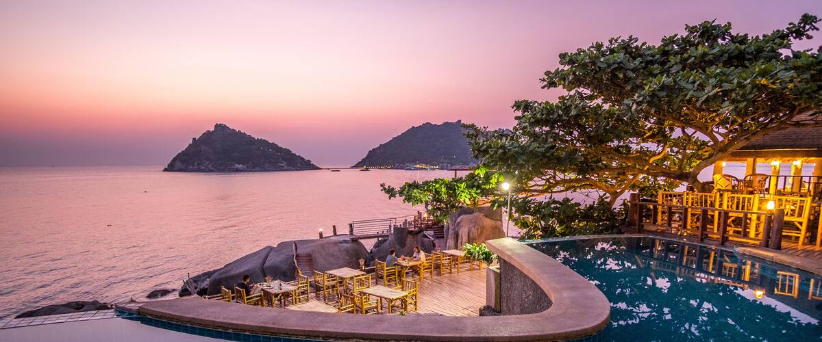 A sunset view of a private pool with a wooden deck and chairs, surrounded by rocks and a tree.