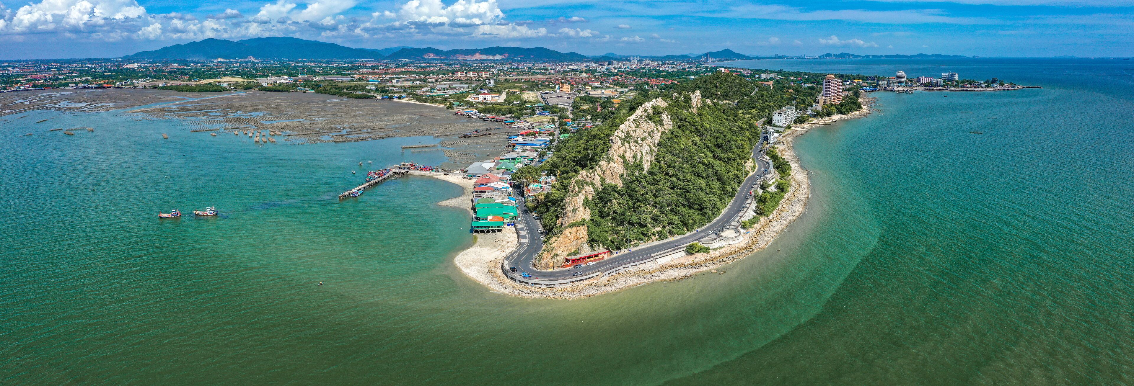 An aerial view of a coastal city with a winding road along the shore, a marina, and a densely populated urban area in the background.