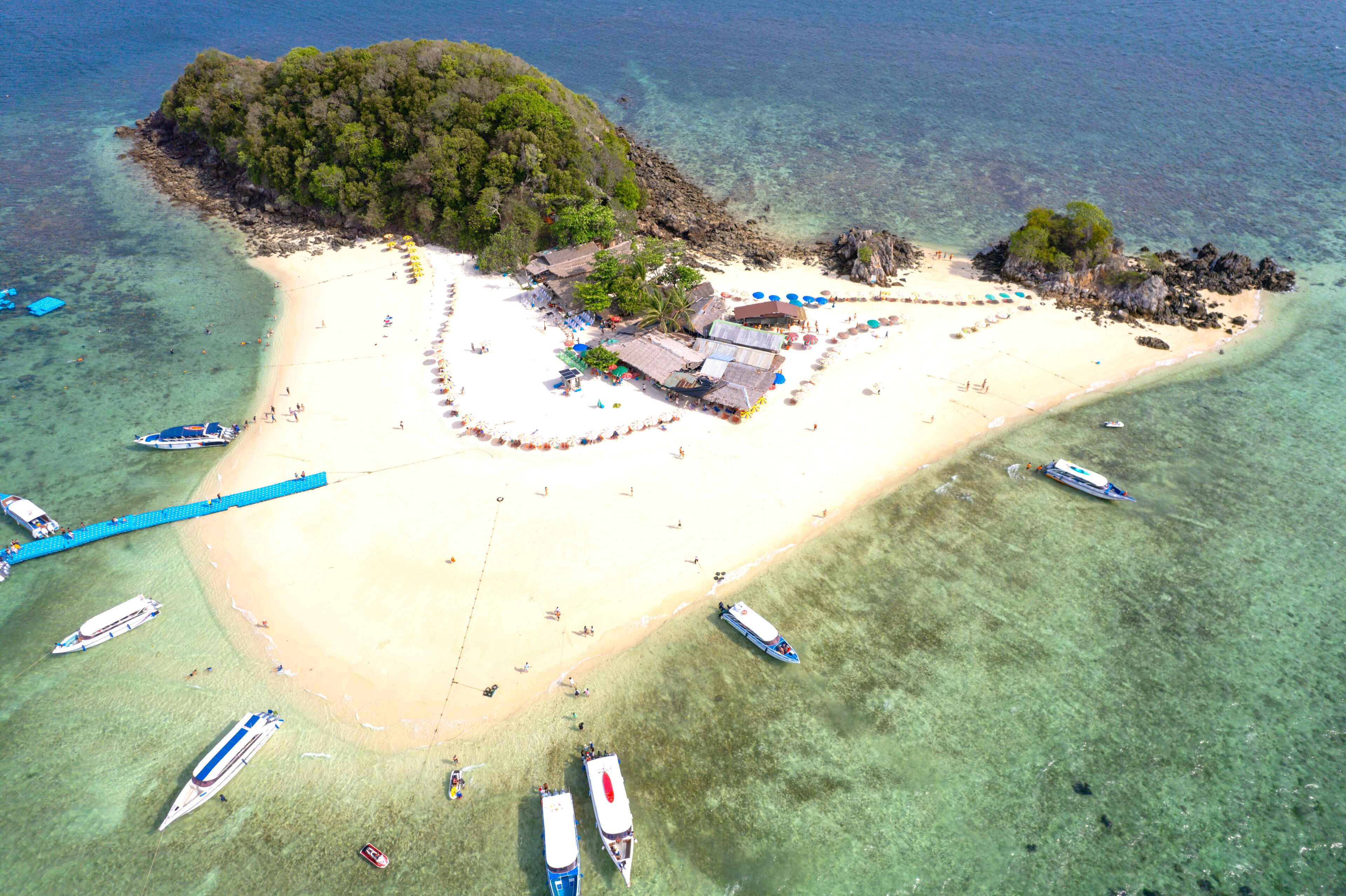 An aerial view of a tropical island with a white sandy beach, clear turquoise waters, and several boats anchored near the shore.