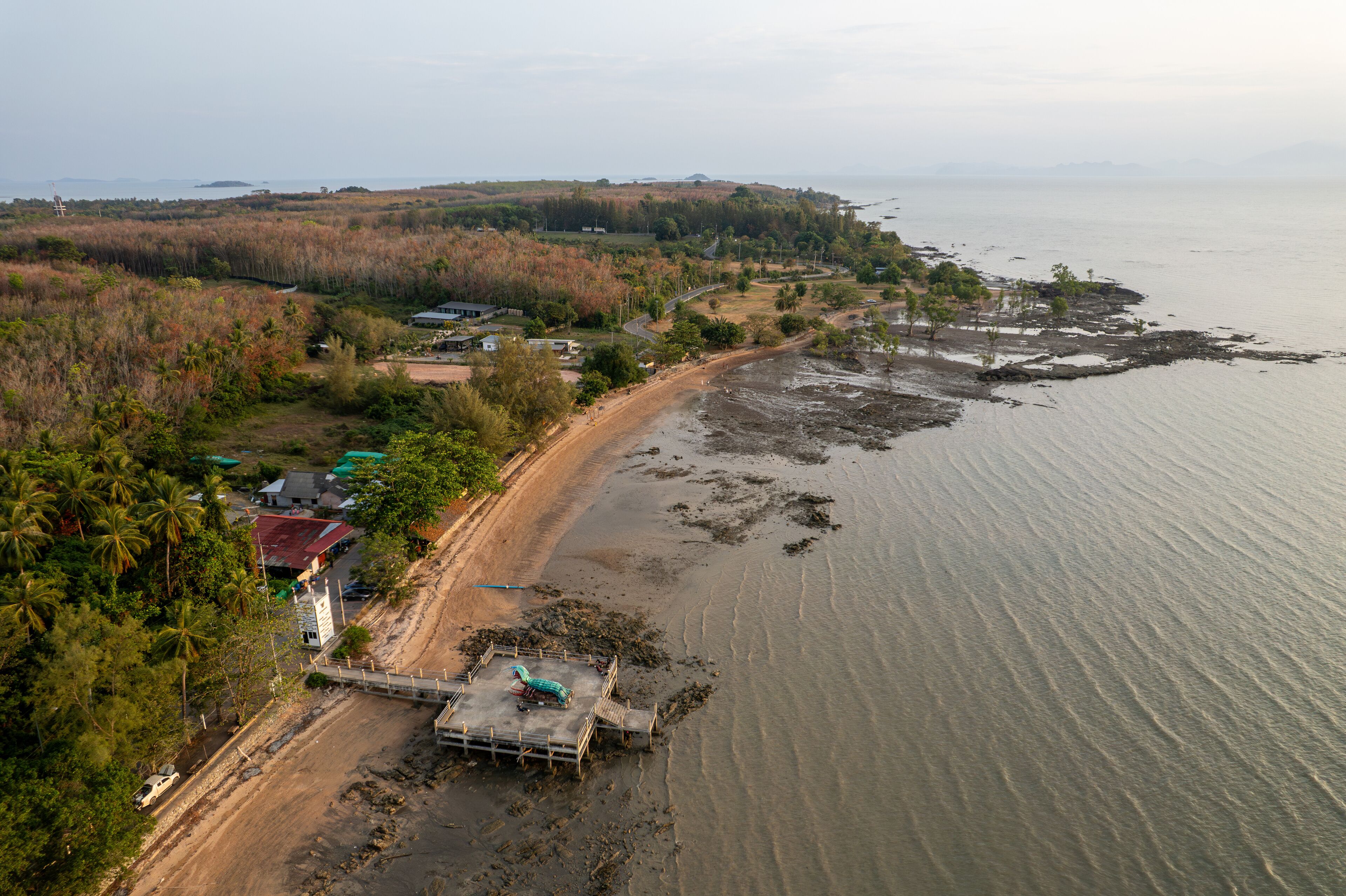 An aerial view of a coastal area with a sandy beach, a small pier, and scattered buildings.