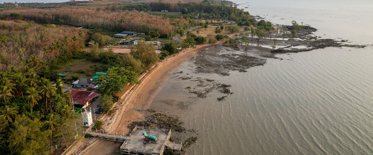 An aerial view of a coastal area with a sandy beach, a small pier, and scattered buildings.