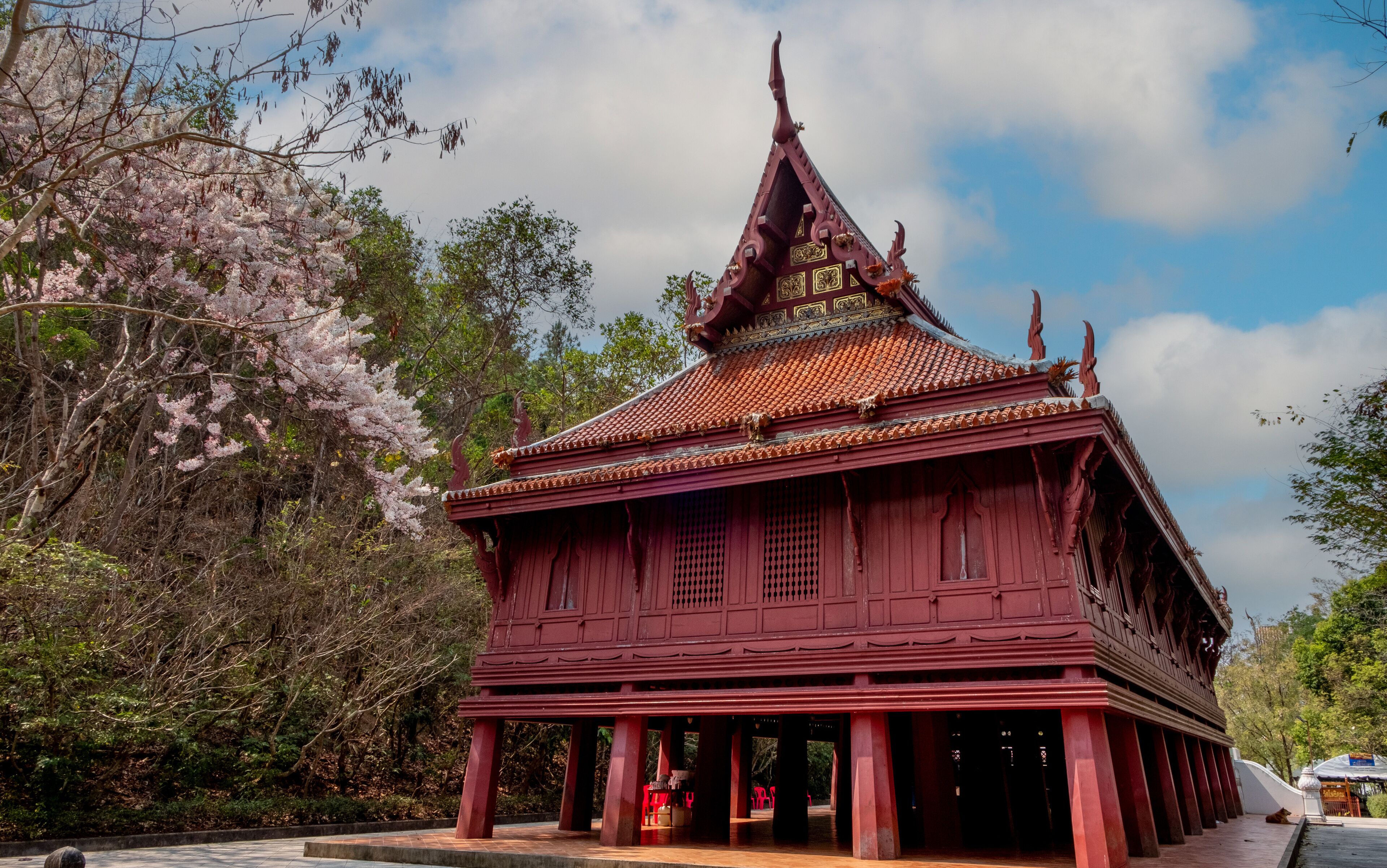A traditional red-roofed temple with ornate decorations and a pointed roof, surrounded by lush greenery and flowering trees.