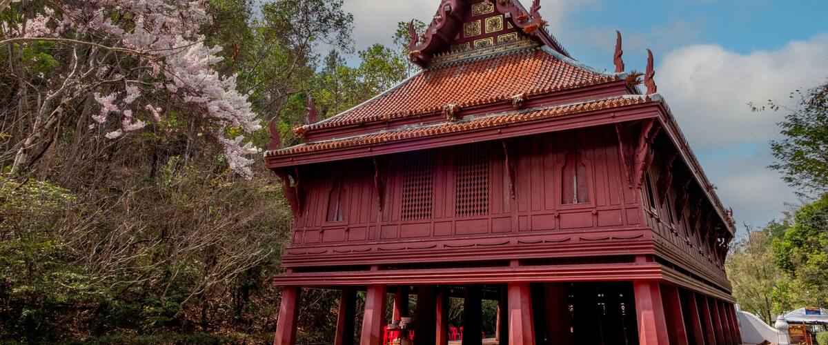 A traditional red-roofed temple with ornate decorations and a pointed roof, surrounded by lush greenery and flowering trees.
